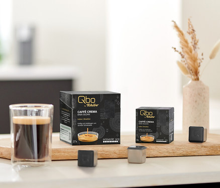 Auf einem Tisch stehen Qbo Caffè BABA BUDAN Kapseln, eine Tasse Kaffee und Kapsel-Prototypen auf einem Holzbrett.