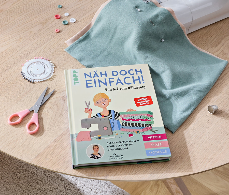 Auf einem Tisch liegen eine Nähmaschine, das Buch »Näh doch einfach«, Stoffe, eine Schere, Knöpfe und Stecknadeln.