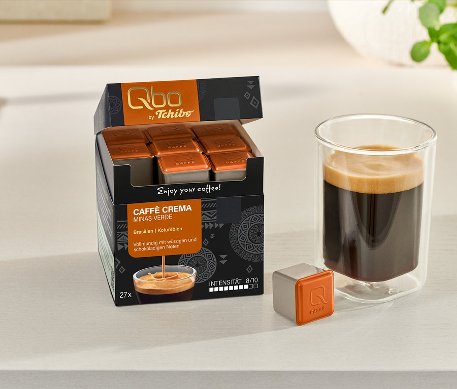 Ein offener Karton Qbo Caffè MINAS VERDE mit Kapseln, eine einzelne Kapsel und ein Glas Kaffee stehen auf einer Theke.