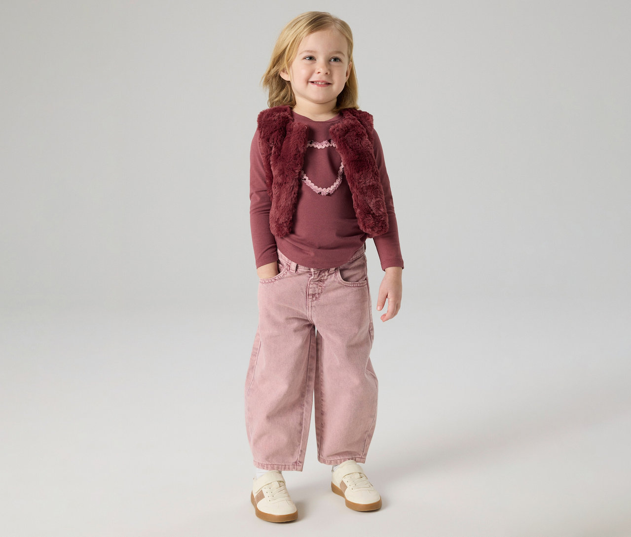 Blondes Mädchen in einem langärmeligen, beerentfarbenen Oberteil, einer beerentfarbenen Kunstfellweste mit hellrosa Herzdetail, weit geschnittenen rosa Hosen mit Händen in den Taschen und hellen Turnschuhen, stehend und lächelnd.