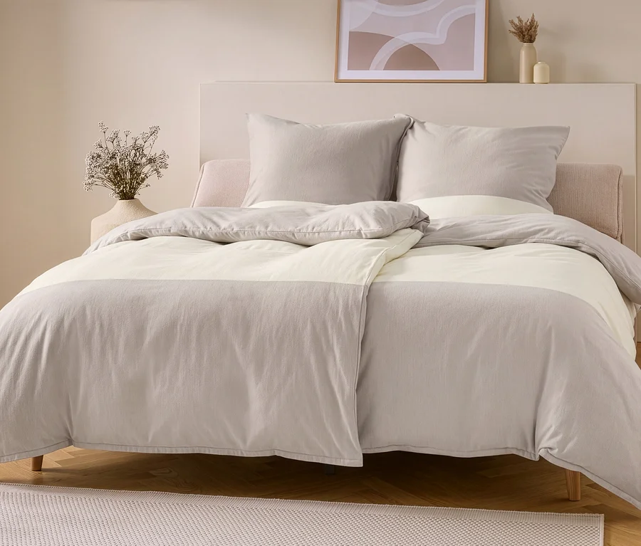 Ein Bett mit taupe-offweißer Flanell-Bettwäsche. Der Bettbezug hat oben und unten taupefarbene Bereiche mit einem offweißen Mittelstreifen, zwei taupefarbene Kissen liegen an einem hellen Kopfteil.