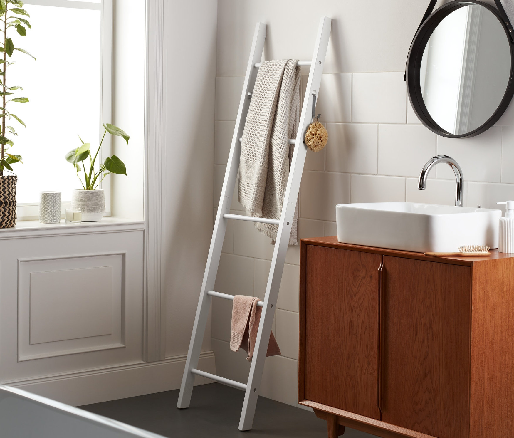Im Badezimmer steht eine weiße Handtuchleiter, an der Handtücher hängen. Daneben befinden sich ein Waschbecken mit braunem Schrank und ein runder Spiegel.