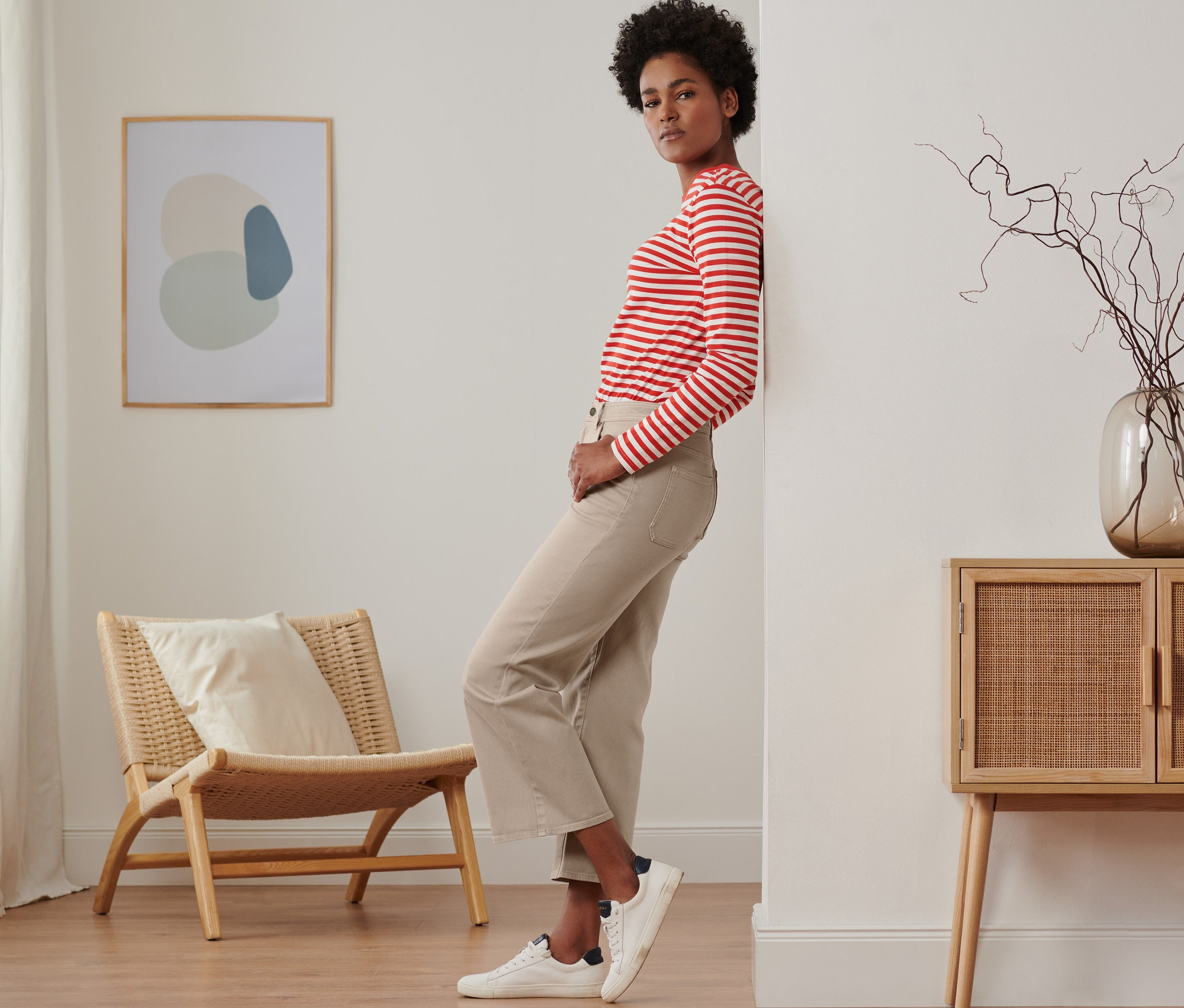 Eine Frau lehnt an einer Wand und trägt Jeans mit weitem Bein, ein rot gestreiftes Oberteil und Turnschuhe.