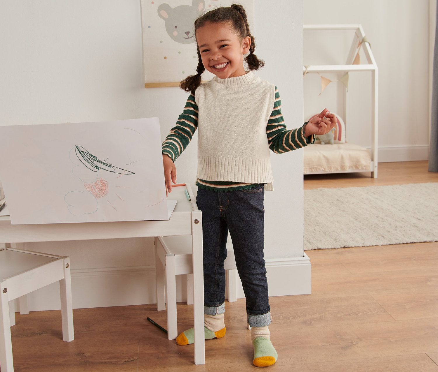 Ein lächelndes Mädchen steht an einem weißen Kindertisch und malt auf ein großes Blatt Papier. Sie trägt einen beigen Strickpullunder mit RWS-Wolle.