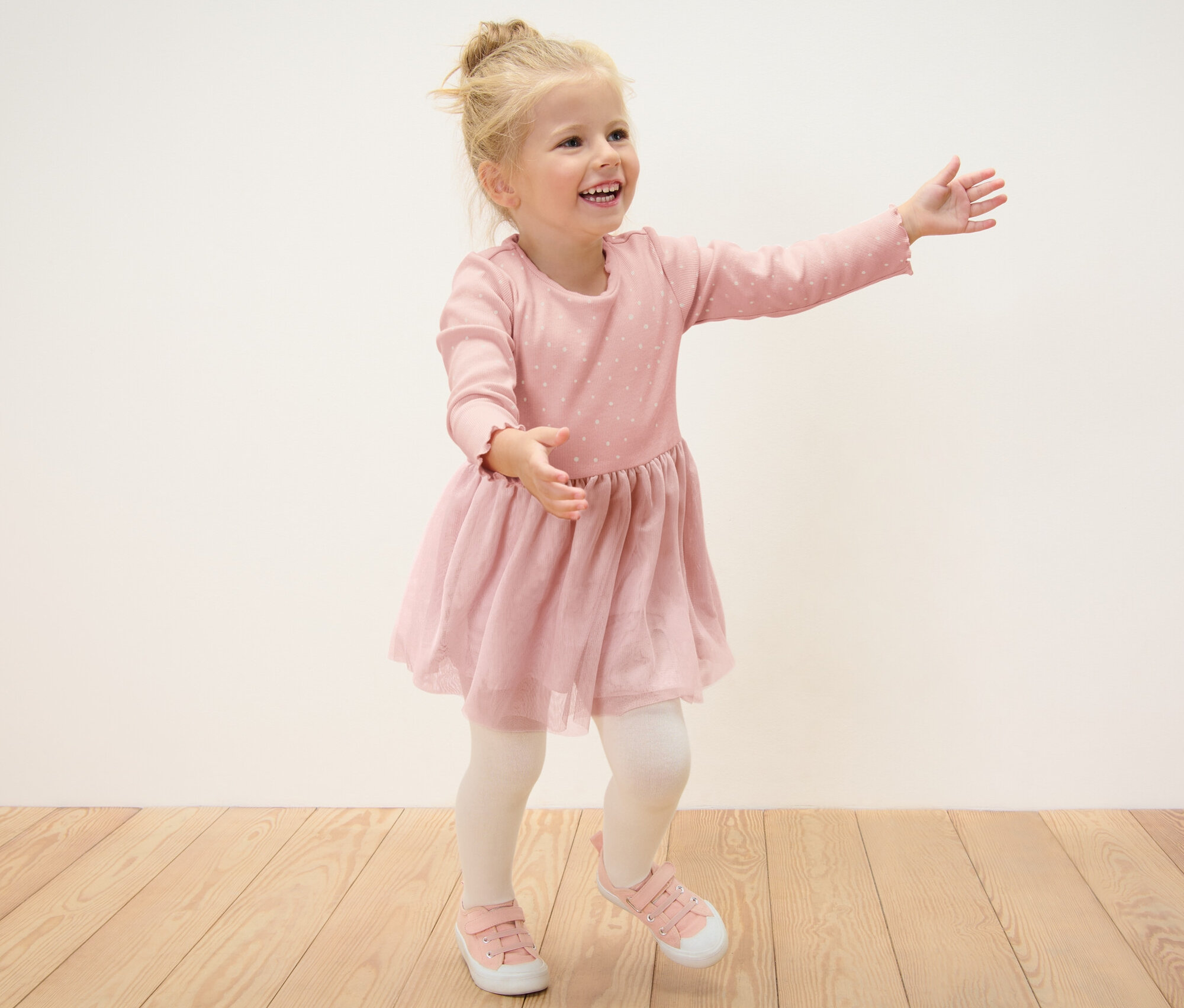 Ein lächelndes blondes Mädchen im Kinder-Rippkleid posiert auf einem Holzboden.