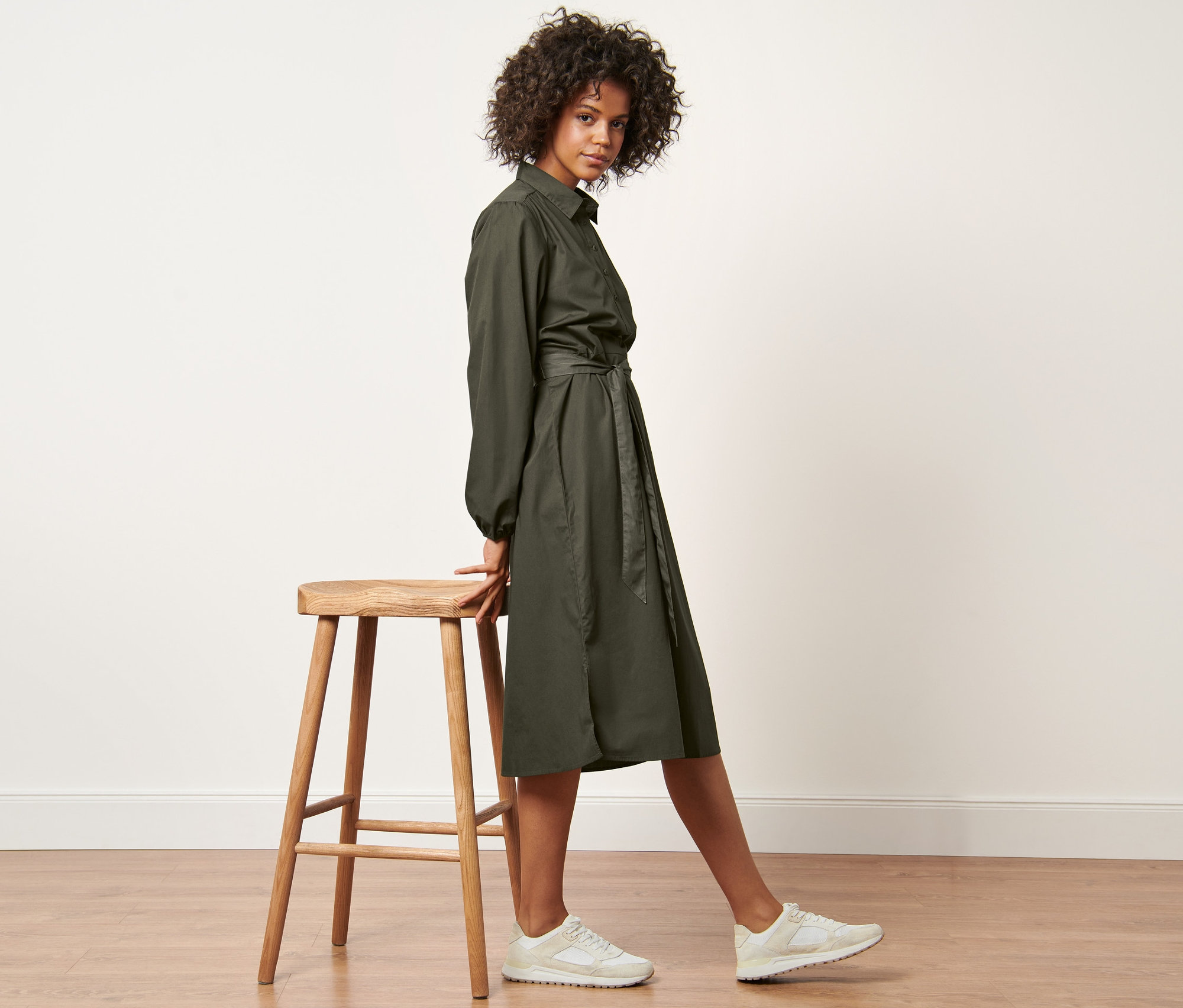 Frau posiert in einem Popeline-Kleid mit Bindegürtel und weißen Turnschuhen neben einem Holzhocker.