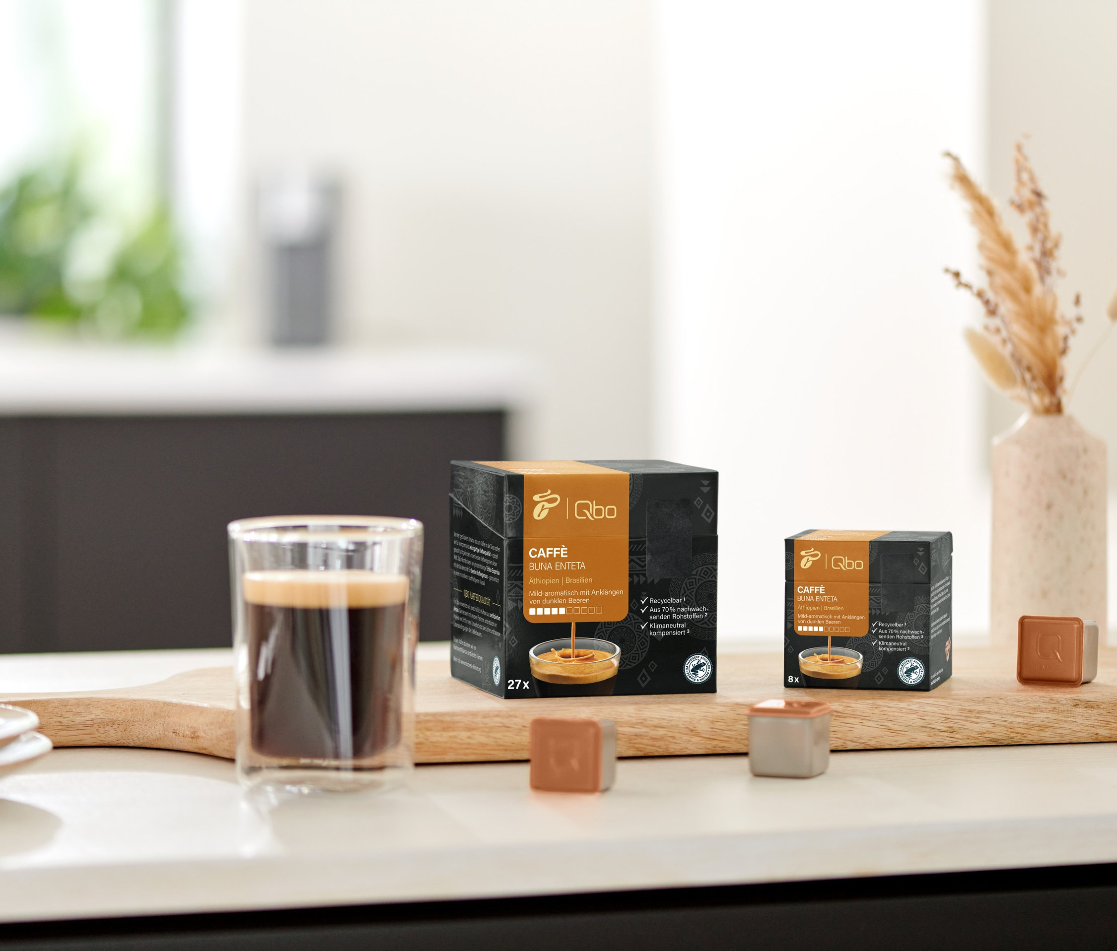 Auf einem Holzbrett stehen ein Glas Kaffee und Packungen von Qbo Caffè BUNA ENTETA.