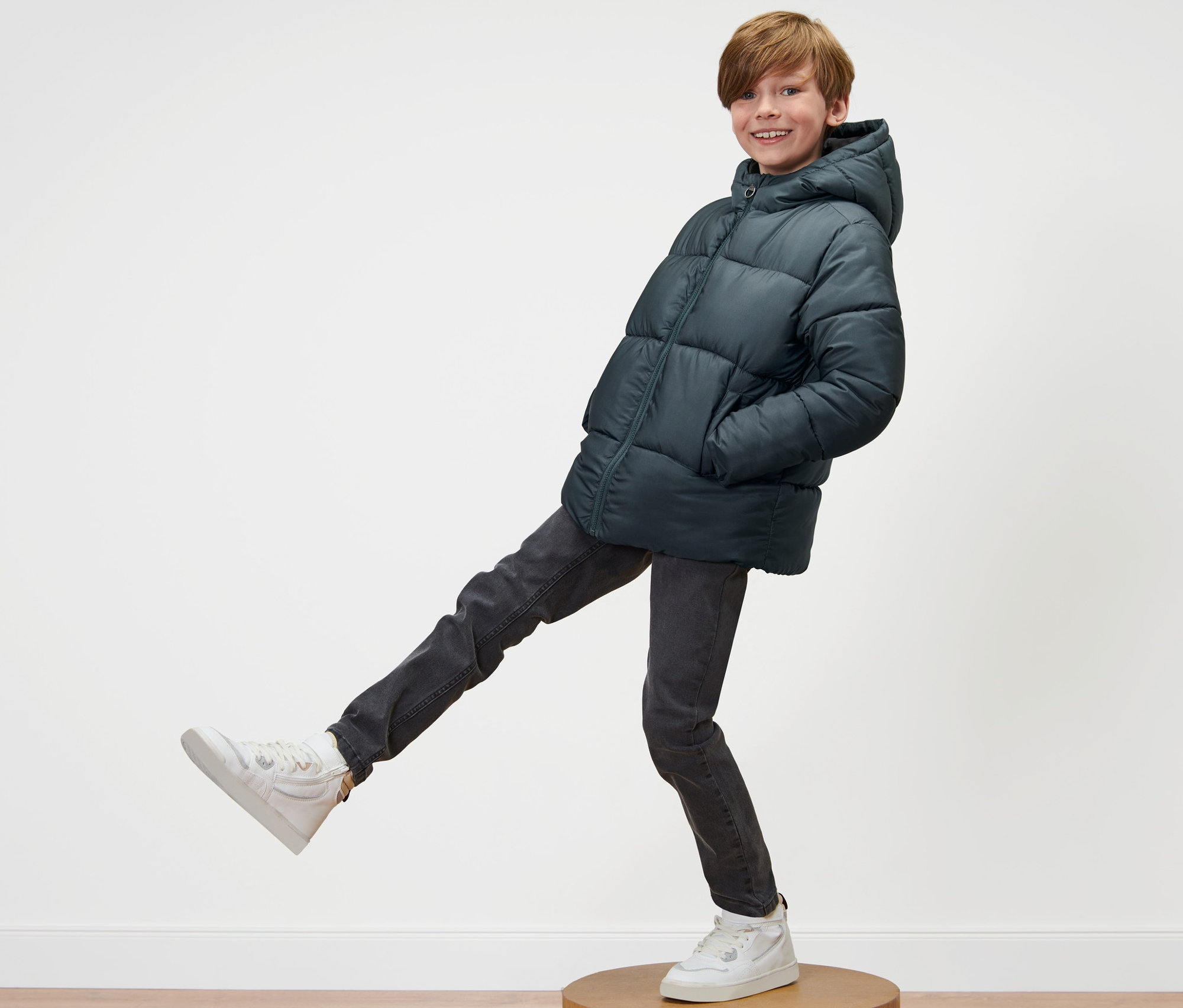 Junge posiert auf einem Holzsockel in Kinder-Thermojacke und Turnschuhen.