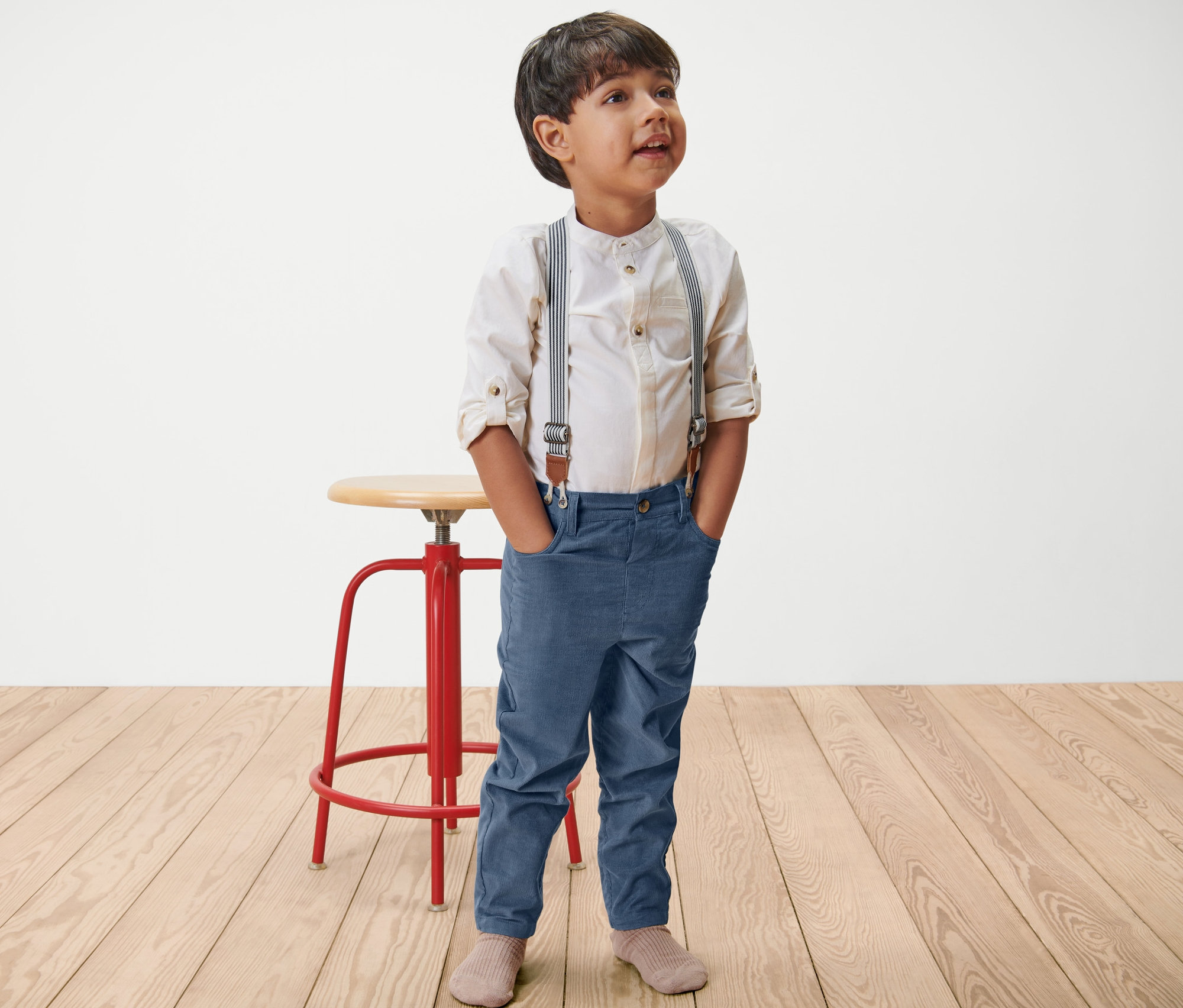 Ein Junge steht mit den Händen in den Taschen und trägt eine Kinder-Cordhose mit abnehmbaren Hosenträgern.