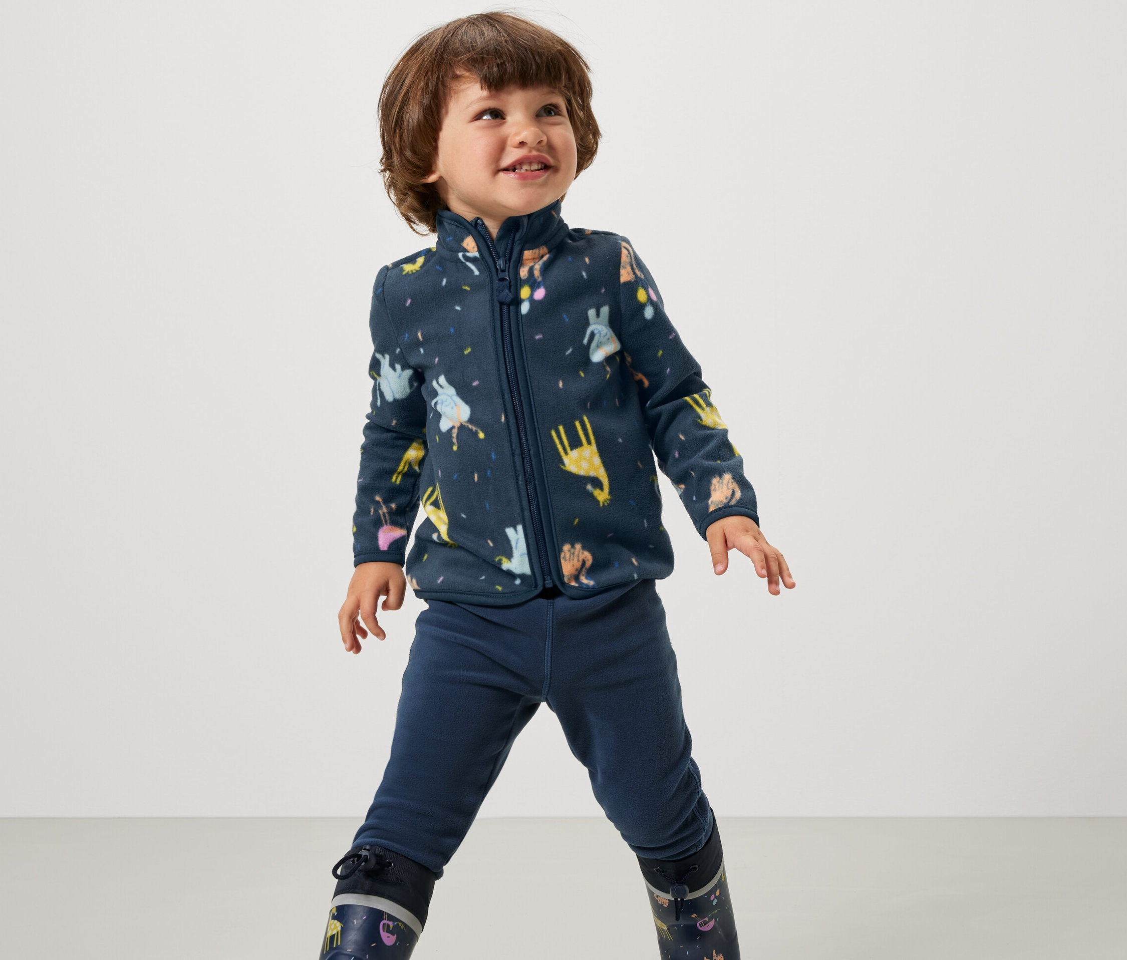 Ein Junge trägt eine Kinder-Fleecejacke mit Tiermuster, blaue Hosen und Gummistiefel mit Tiermuster.
