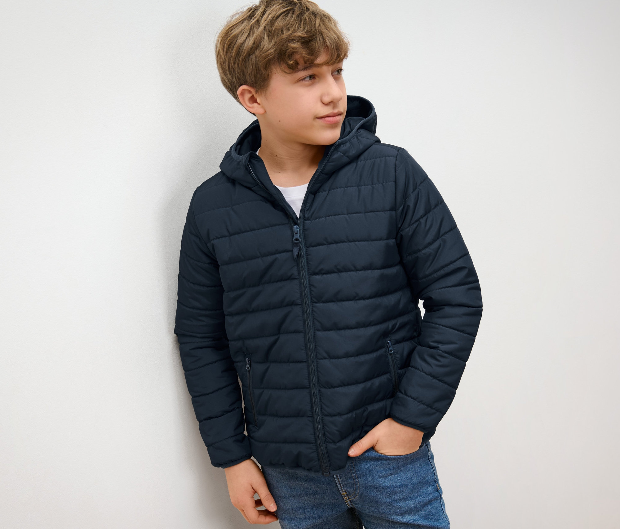 Junge in blauer Kinder-Steppjacke steht an einer Wand.
