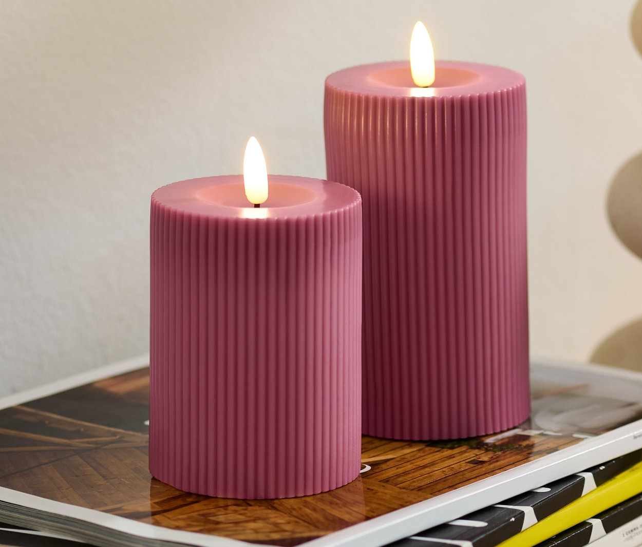 Zwei rosafarbene LED-Echtwachskerzen stehen auf einem Stapel Zeitschriften. Im Hintergrund eine Vase mit Eukalyptus.