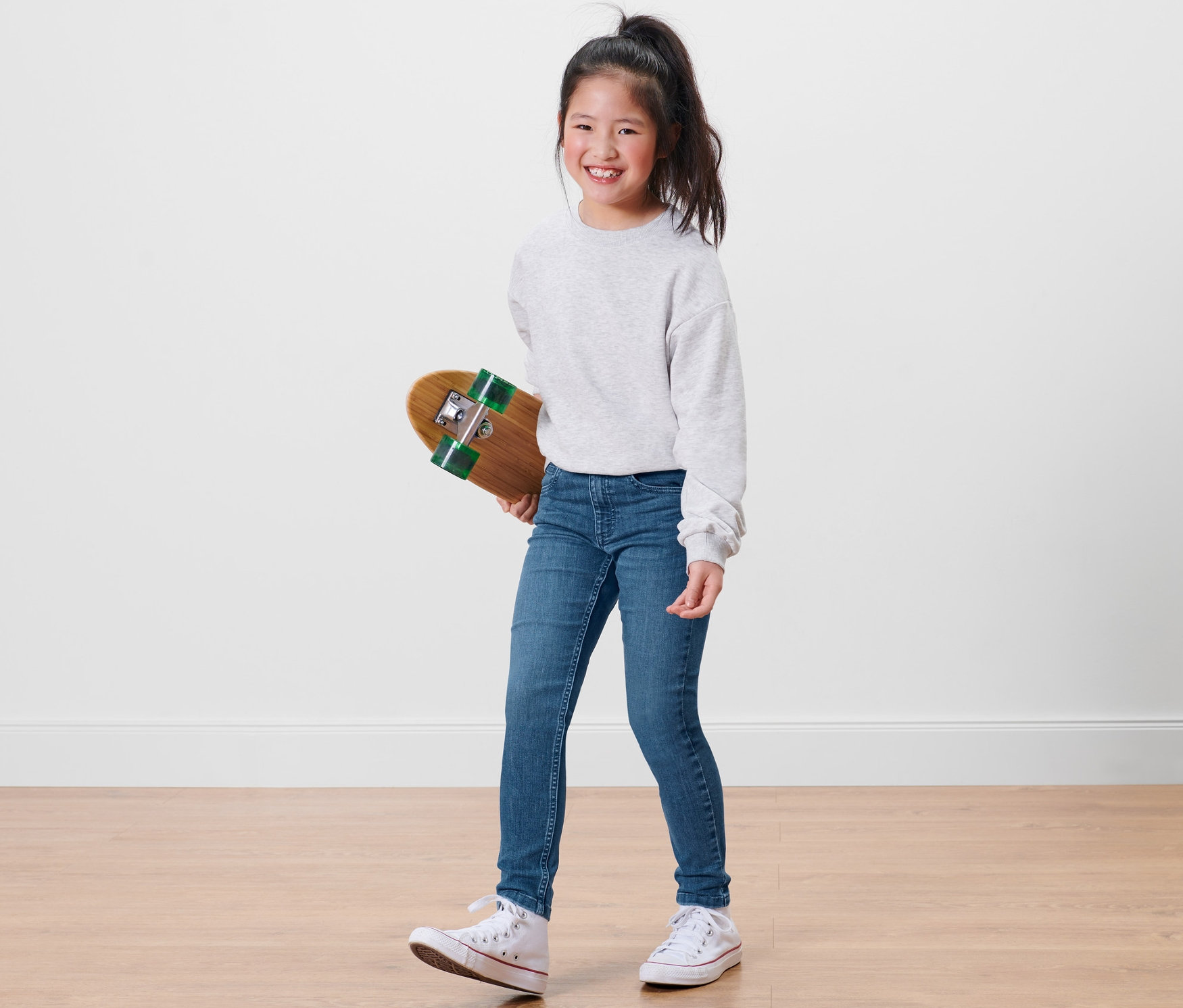 Lächelndes Mädchen hält ein Skateboard und trägt eine blaue Kinder-High-Waist-Jeans – Fit »Lotta«.