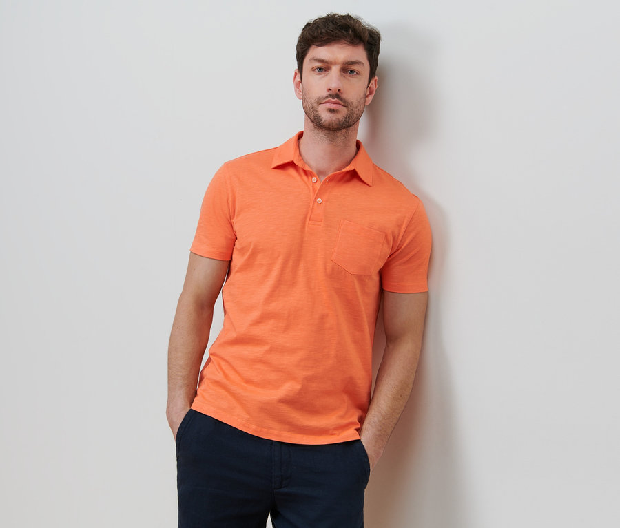 Mann lehnt an Wand, trägt orangefarbenes Jersey-Poloshirt.