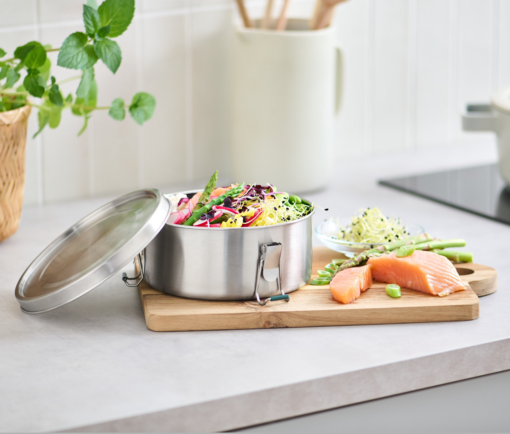 Eine Edelstahl-Lunchbox gefüllt mit Gemüse und Salat, daneben Lachs und Spargel auf einem Schneidebrett.