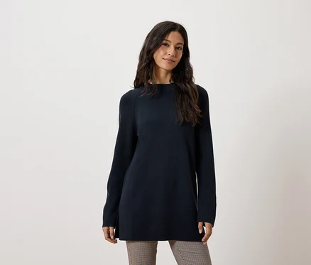Eine Frau trägt einen langen, dunkelblauen Pullover und eine braune Hose.