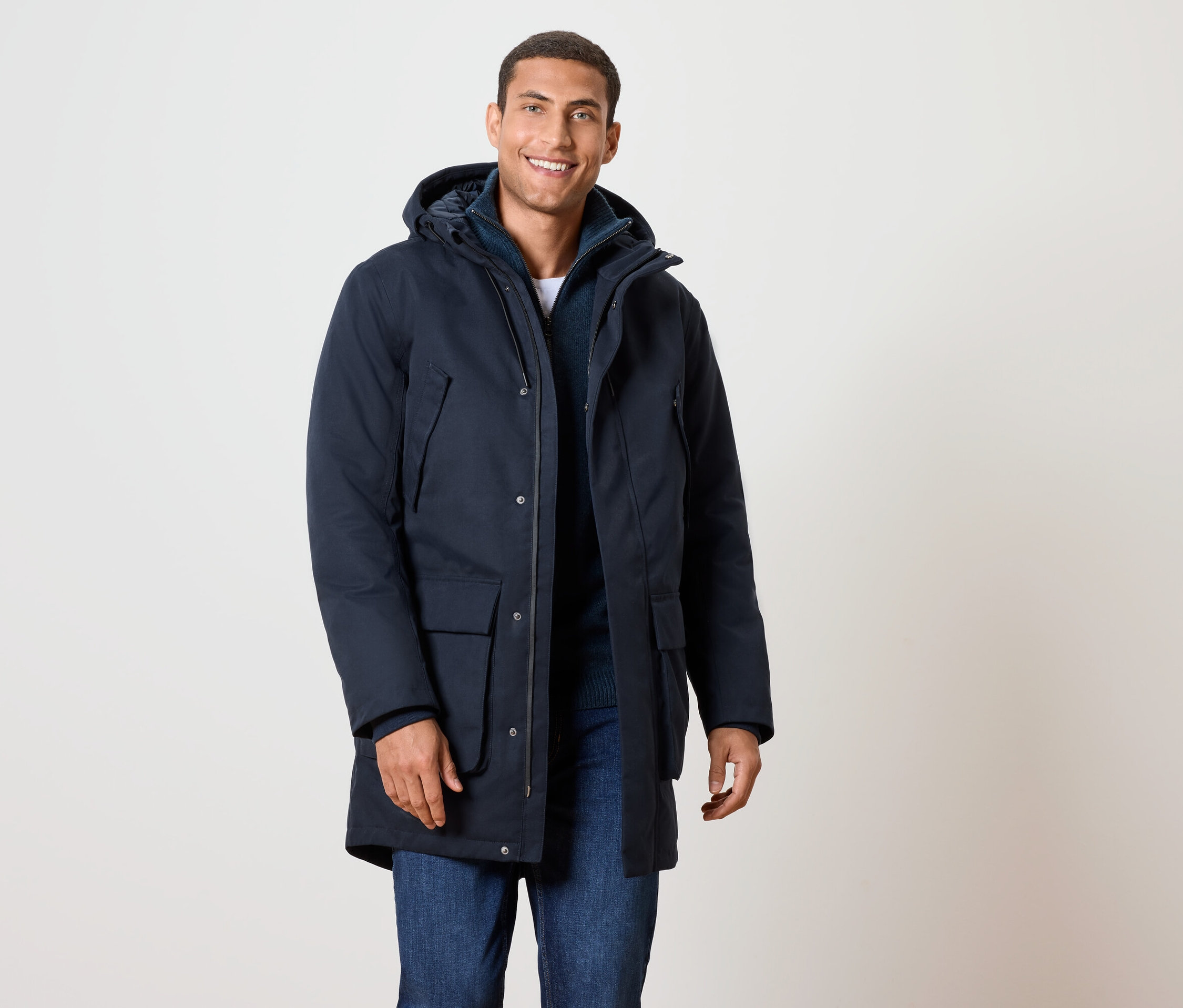 Ein lächelnder Mann trägt eine dunkelblaue Winterjacke mit Kapuze und Jeans.