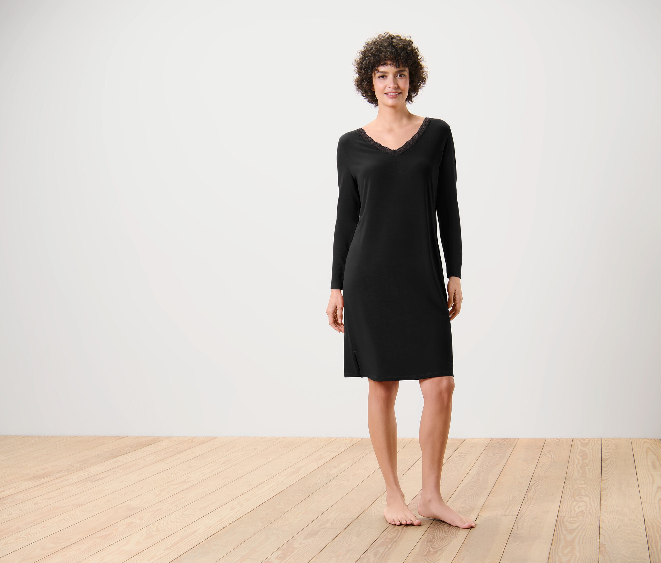 Eine Frau steht barfuß auf einem Holzboden und trägt ein schwarzes, langärmliges Kleid.