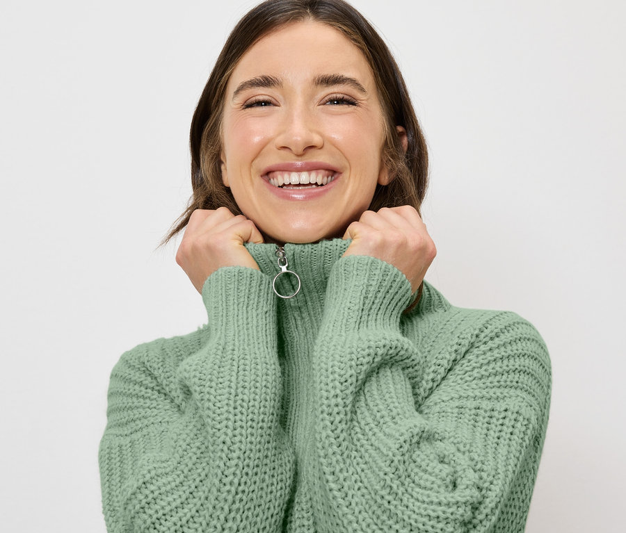 Lächelnde Frau hält den Kragen eines hellgrünen Pullovers mit Reißverschluss mit ihren Händen.