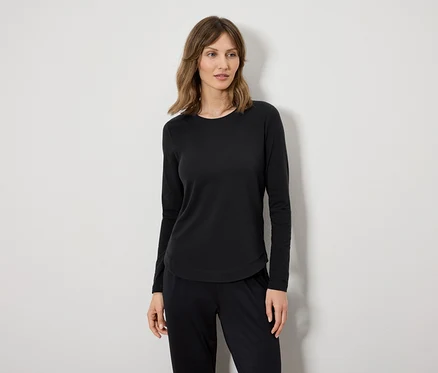 Eine Frau in einem schwarzen Langarmshirt und einer schwarzen Hose steht vor einer weißen Wand.