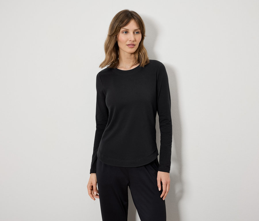 Eine Frau in einem schwarzen Langarmshirt und einer schwarzen Hose steht vor einer weißen Wand.