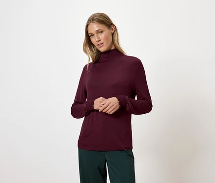 Blonde Frau trägt ein weinrotes Rollkragenshirt und grüne Hosen.