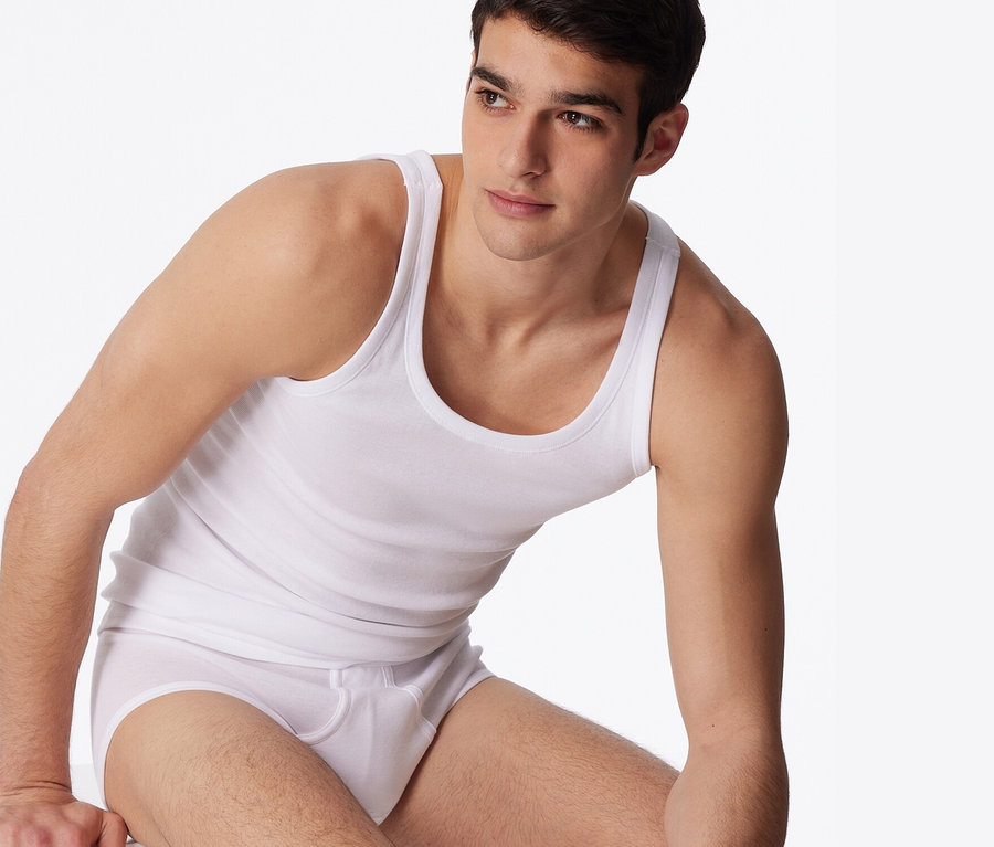 Mann sitzt vor einem weißen Hintergrund und trägt 2 weiße SCHIESSER Unterhemden in Feinripp.