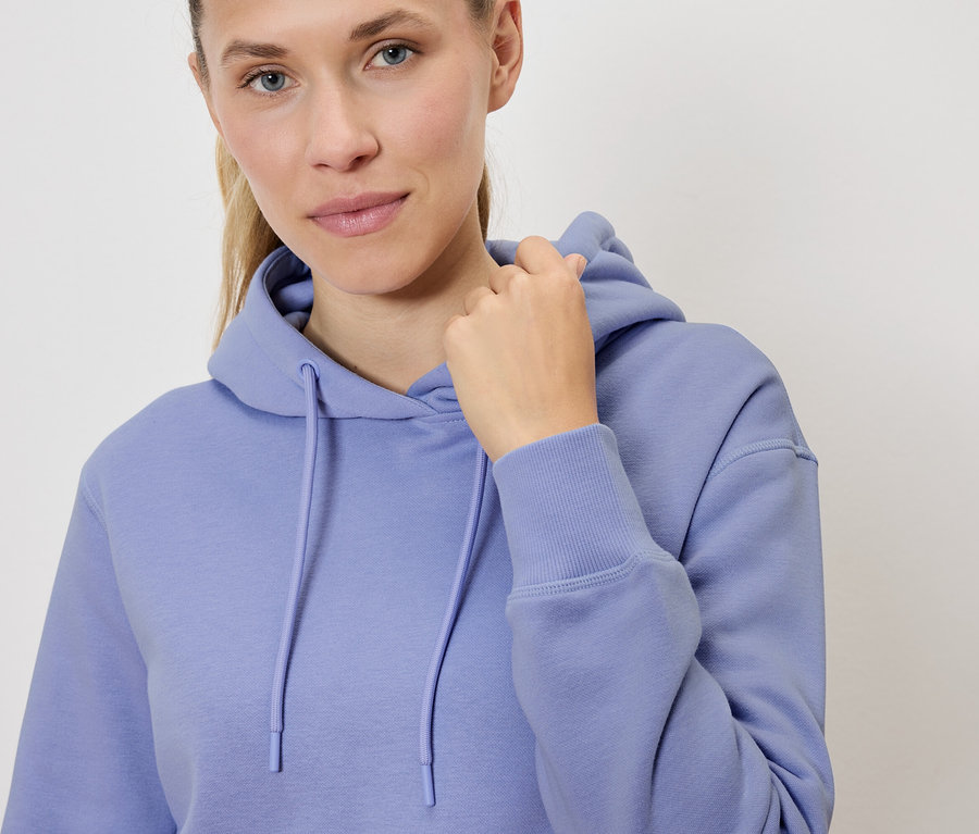 Porträt einer blonden Frau in einem lila Kapuzenpullover, die ihre Kapuze festhält.