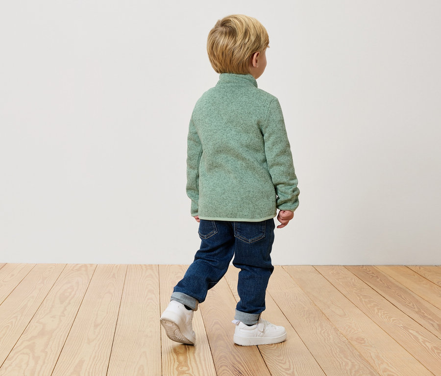 Ein Kind geht mit dem Rücken zur Kamera in einem grünen Fleecepullover, Jeans und weißen Turnschuhen auf einem Holzboden.