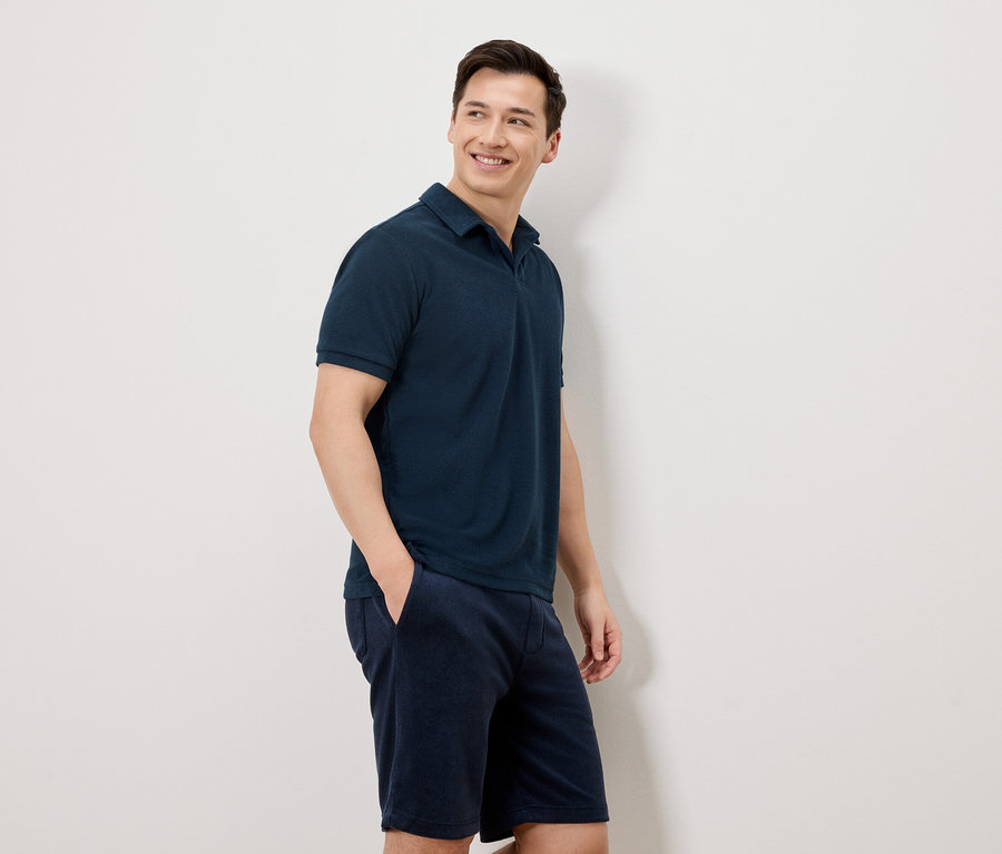 Ein Mann mit dunklen Haaren, der ein dunkelblaues Poloshirt und Shorts trägt, steht vor einer weißen Wand.