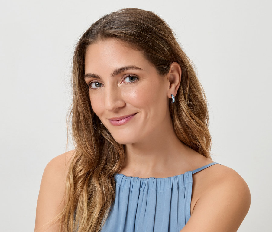 Frau mit langen, gewellten, braunen Haaren und blauem Oberteil, die blaue Ohrringe trägt und in die Kamera lächelt.