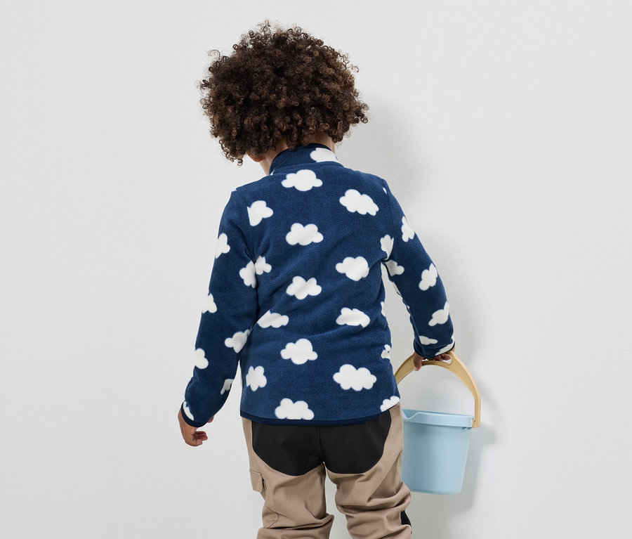 Ein Kind mit lockigem Haar trägt ein blaues Fleeceoberteil mit Wolkenmuster und eine beige Hose und hält einen blauen Eimer.