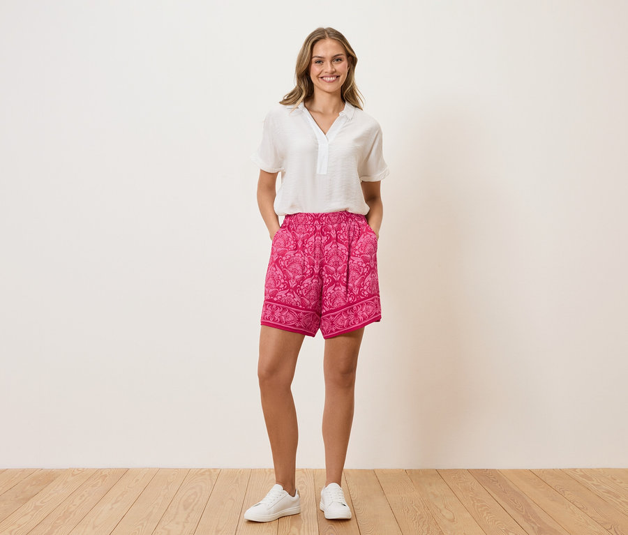 Eine Frau steht mit ihren Händen in den Hosentaschen vor einer Wand. Sie trägt eine weiße Bluse, pinke Shorts mit Muster und weiße Turnschuhe.