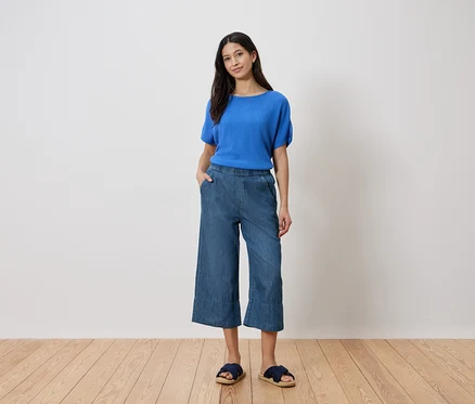 Frau steht mit Hand in der Hosentasche, trägt blaues Oberteil, Jeans-Culottes und Sandalen vor einer weißen Wand.