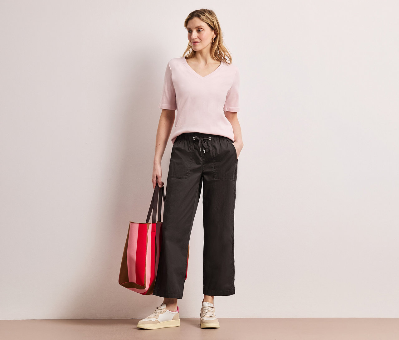 Frau mit rosa Oberteil und Street One Papertouch-Hose posiert mit einer rot gestreiften Tasche.