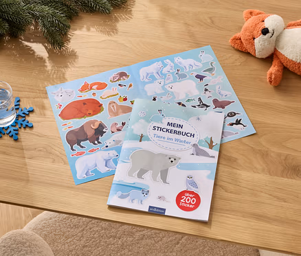 Auf einem Holztisch liegen ein Glas Wasser auf einem blauen Schneeflocken-Untersetzer, ein geöffnetes Stickerbuch mit Wintertiermotiven und ein orangefarbenes Fuchs-Plüschtier.