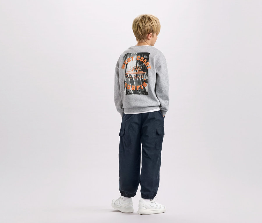 Ein Kind von hinten, trägt einen grauen Pullover mit "West Coast Surfing"-Aufdruck, marineblaue Cargohosen und weiße Turnschuhe.