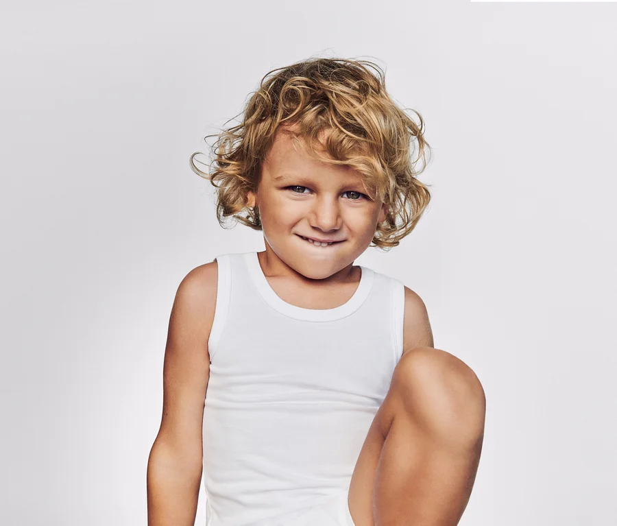 Kind mit blonden Locken und weißem Unterhemd lächelt verschmitzt.