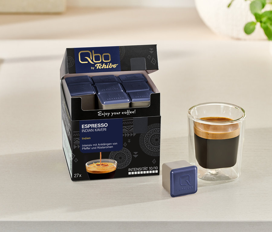 Eine geöffnete Packung Qbo Espresso INDIAN KAVERI mit Kapseln, eine einzelne Kapsel und ein Glas Espresso stehen auf einer hellen Oberfläche.