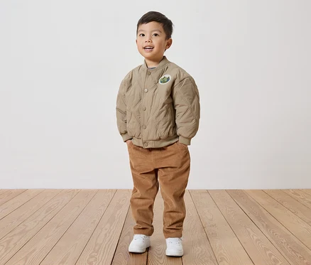 Ein lächelnder Junge mit beiger, gesteppter Jacke mit Aufnäher, Cordhose und weißen Schuhen steht auf einem Holzboden vor einer weißen Wand.