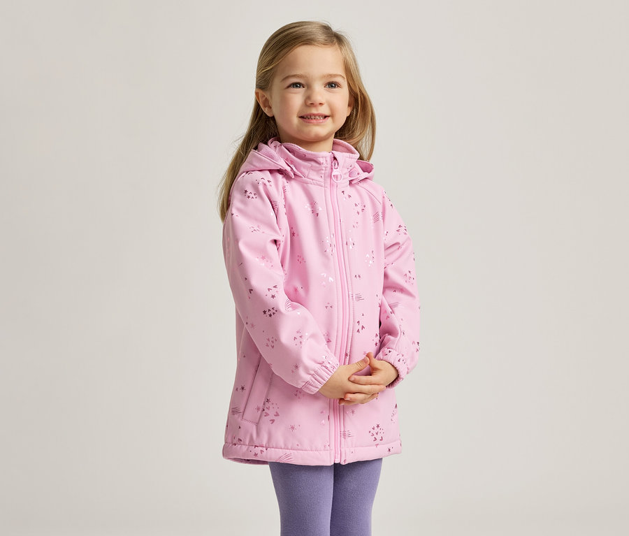 Ein junges Mädchen mit blonden Haaren lächelt und trägt eine rosa, gemusterte Jacke und lila Leggings.