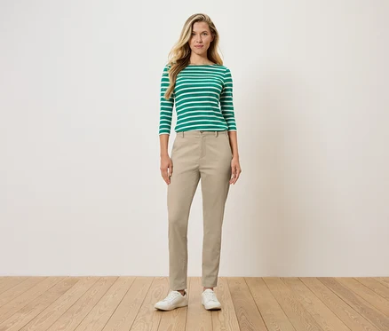 Frau steht und trägt ein grün-weiß gestreiftes Langarmshirt, eine beige Hose und weiße Schuhe.