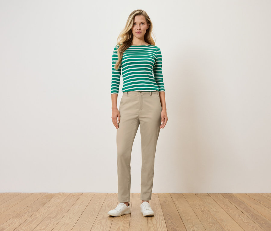 Frau steht und trägt ein grün-weiß gestreiftes Langarmshirt, eine beige Hose und weiße Schuhe.