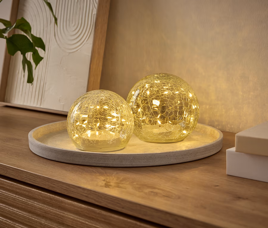 Zwei leuchtende, kugelförmige Dekoleuchten mit Craquelé-Effekt, gefüllt mit kleinen LEDs, stehen auf einem runden Tablett auf einer Holzkommode. Eine Pflanze und ein gerahmtes Bild sind im Hintergrund.