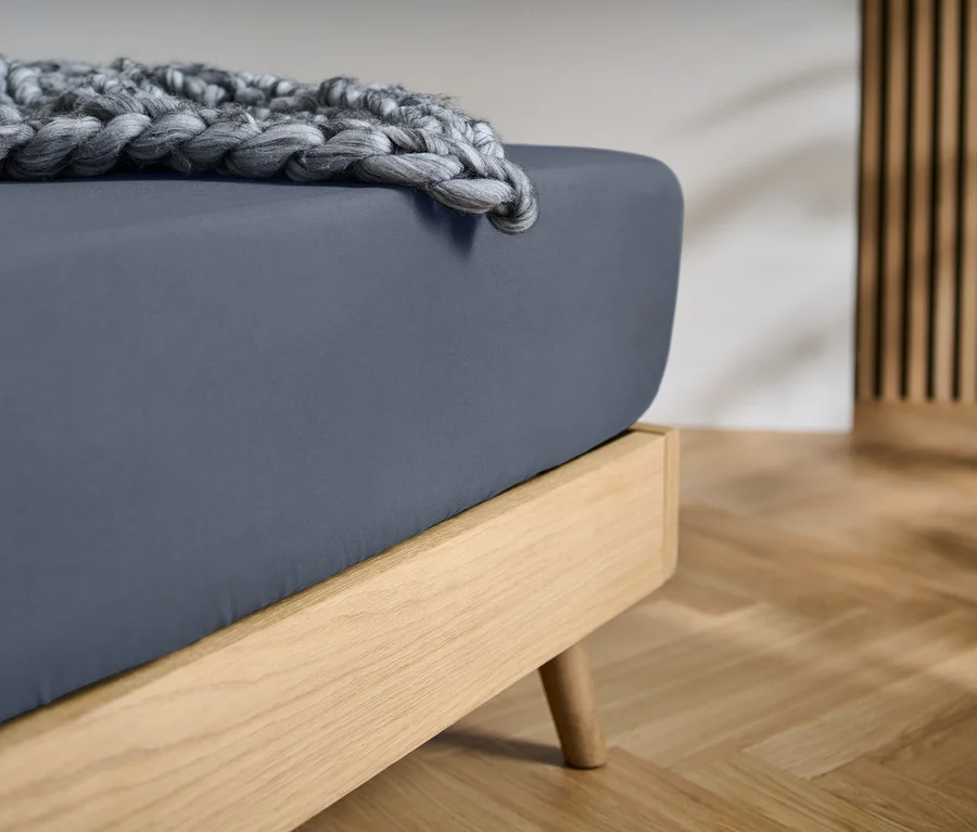 Ein Bett mit hellem Holzrahmen, einem dunkelgrauen Spannbetttuch und einer grob gestrickten grauen Decke. Im Hintergrund sind eine Holzlamellenwand und ein heller Holzfußboden zu sehen.