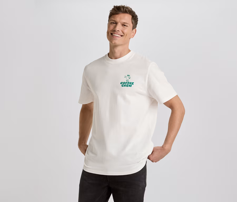 Mann lächelt und posiert in einem cremefarbenen T-Shirt mit dem grünen Aufdruck "Coffee Crew" und schwarzen Jeans vor einem weißen Hintergrund.