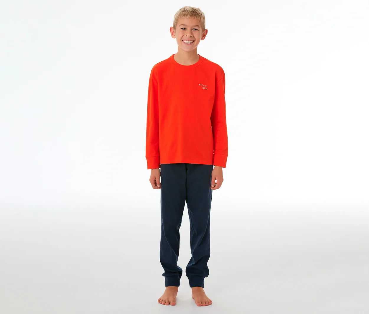 Lächelnder Junge in einem orangefarbenen SCHIESSER Kinder-Pyjama mit Bündchen und dunkelblauer Hose, barfuß stehend.