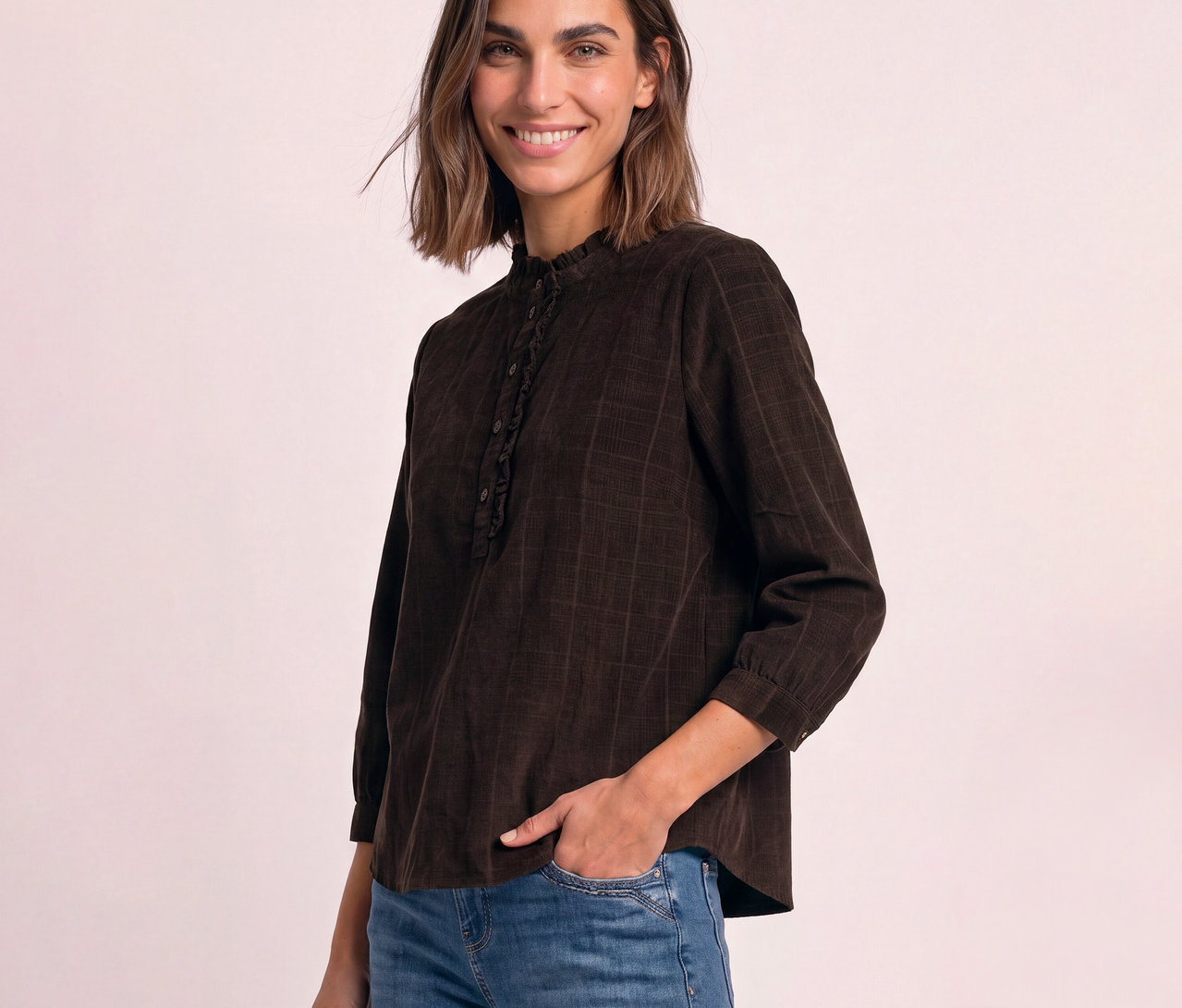 Lächelnde Frau in brauner Street One Cordbluse mit Stehkragen und Rüschen, die linke Hand in der Hosentasche.