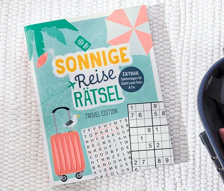 Buch »Sonnige Reise Rätsel« liegt neben einem offenen Koffer.