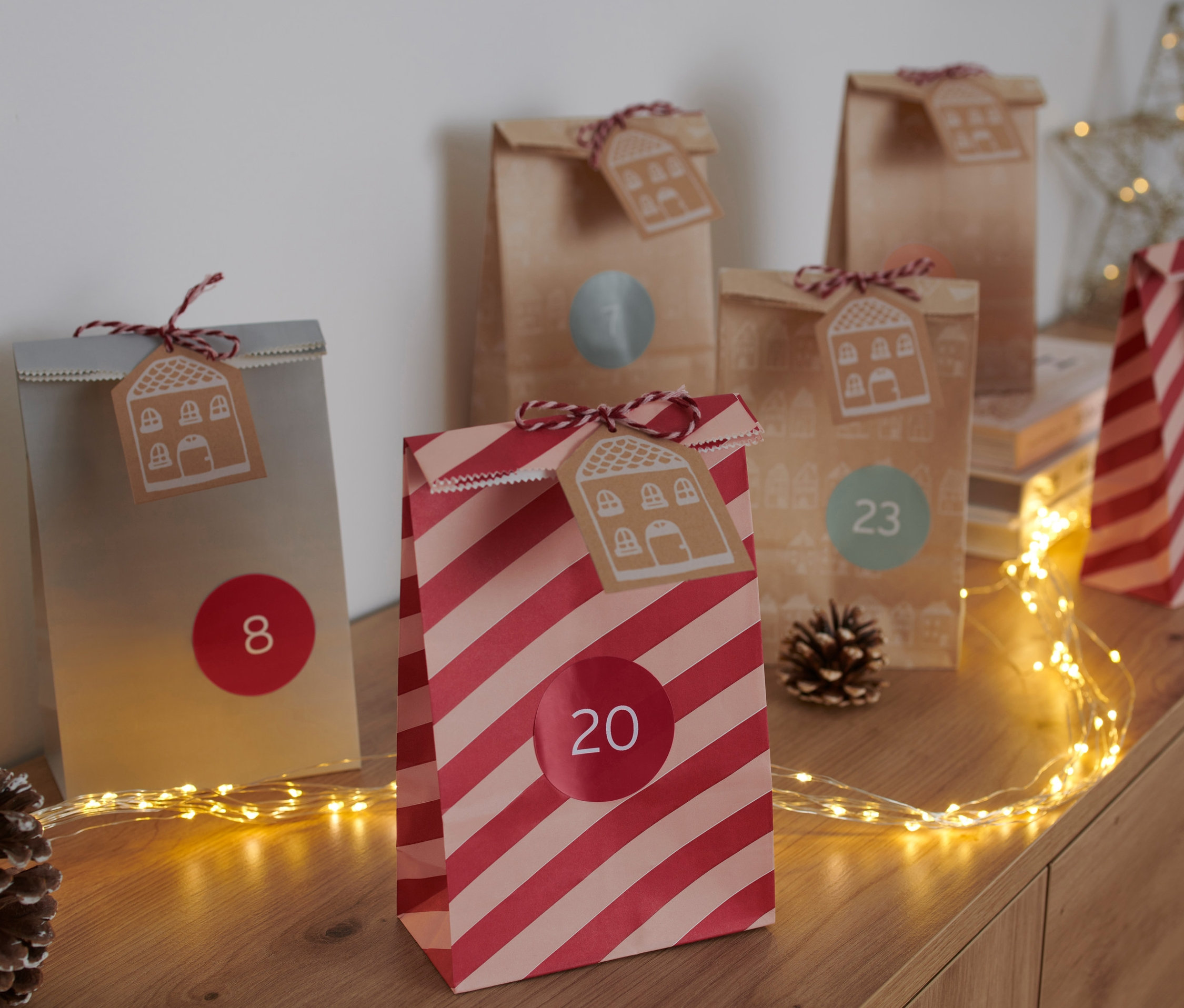 Adventskalender-Tüten-Set mit den Nummern 8, 20 und 23 auf einem Tisch mit Tannenzapfen und Lichtern.
